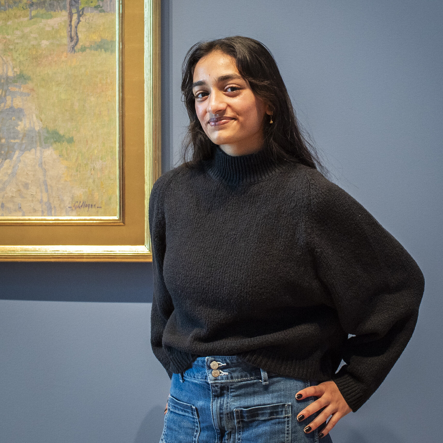 Young woman with long black hair standing with hands on hips in a black sweater in front of an Impressionist artwork and blue wall in an art museum