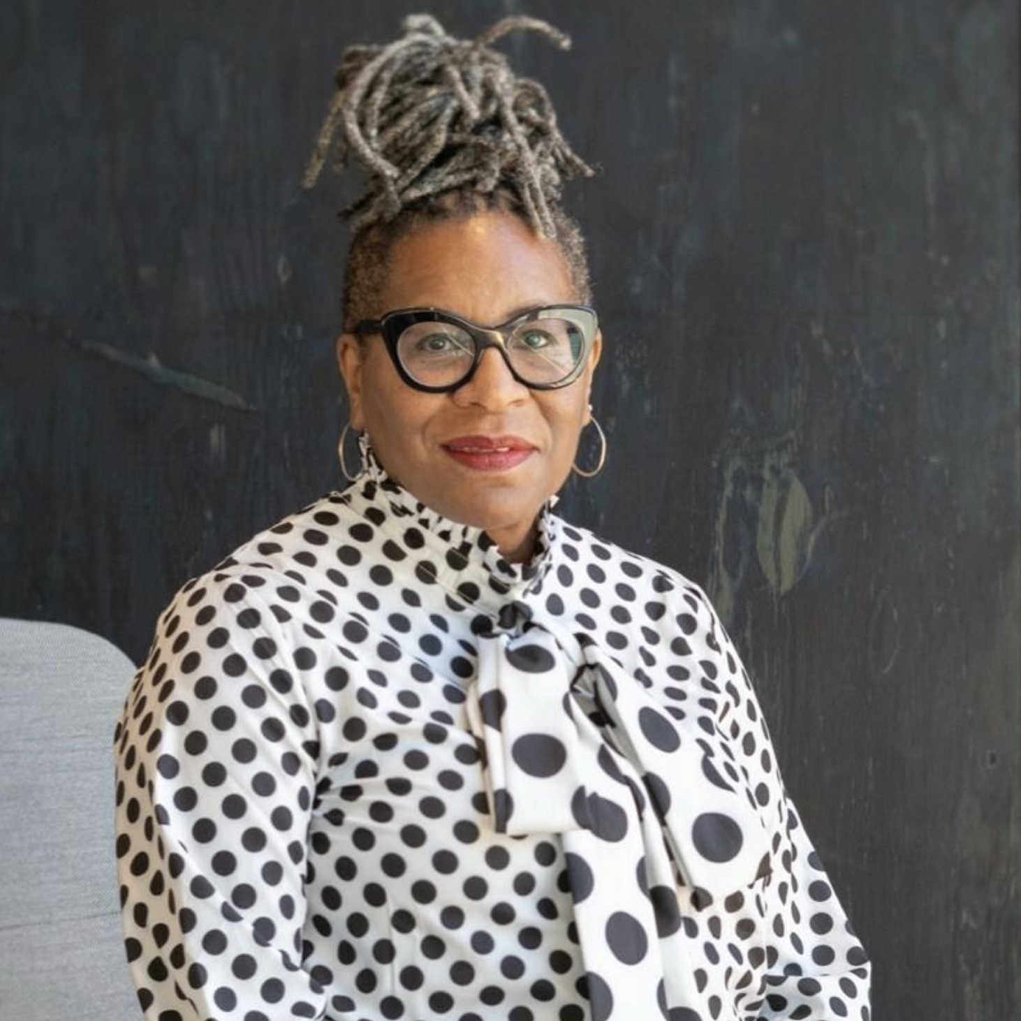 Woman in a black and white spotted blouse in front of a grey wall with glasses, red lipstick, and grey locks