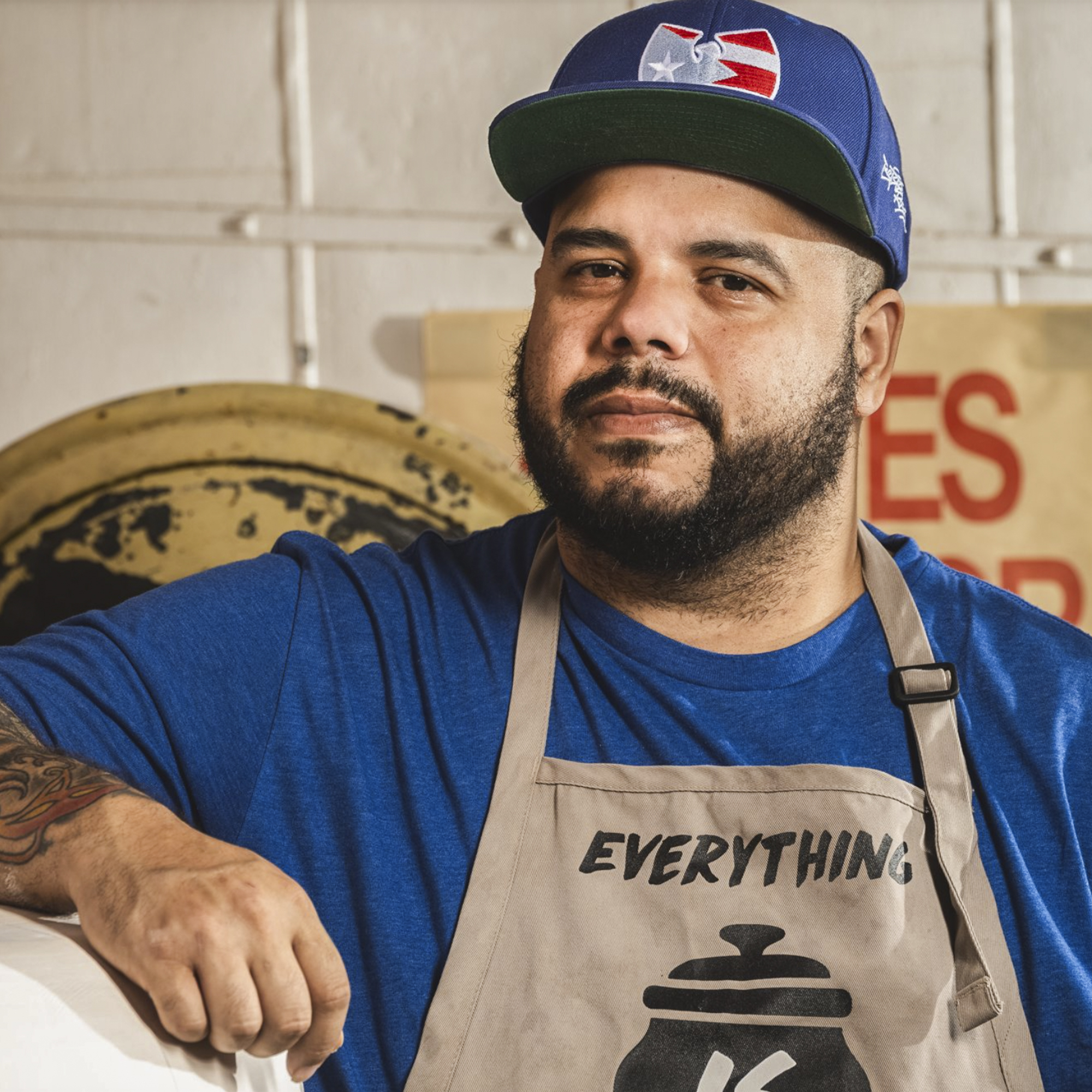 Image of a bearded man with a ball cap, blue shirt, and tan apron resting his elbow on a piece of pottery