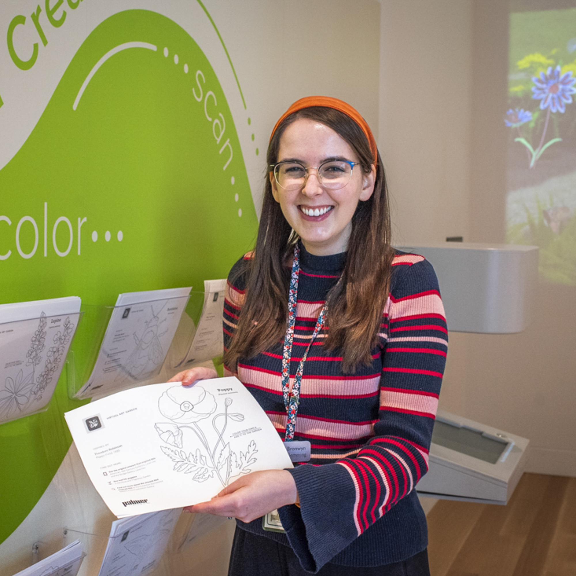 A woman in a pink, red, and blue striped sweater and orange headband with glasses holding a worksheet in front of a green wall