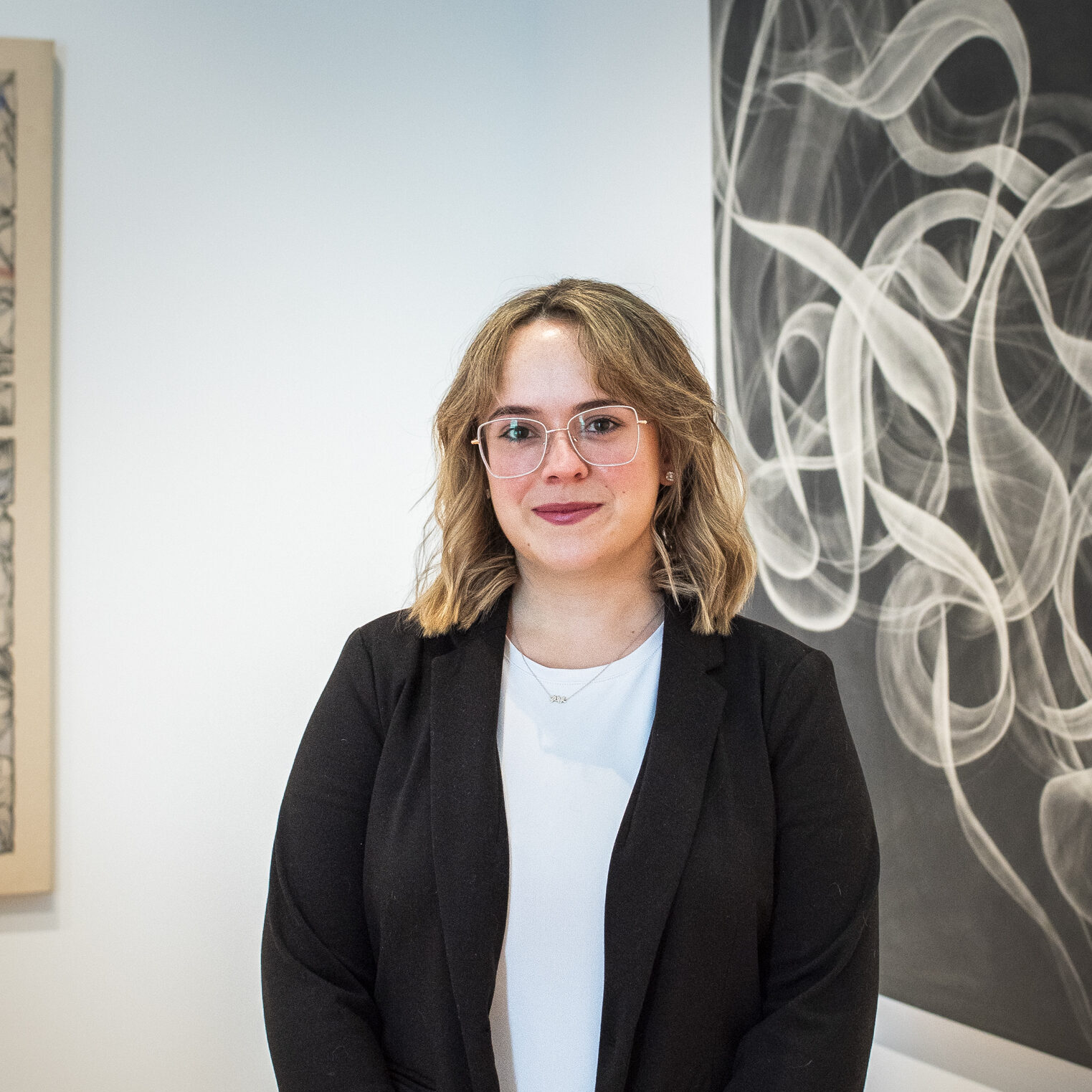 A woman in a black blazer and white shirt standing with hands clasped in front of two black and white artworks