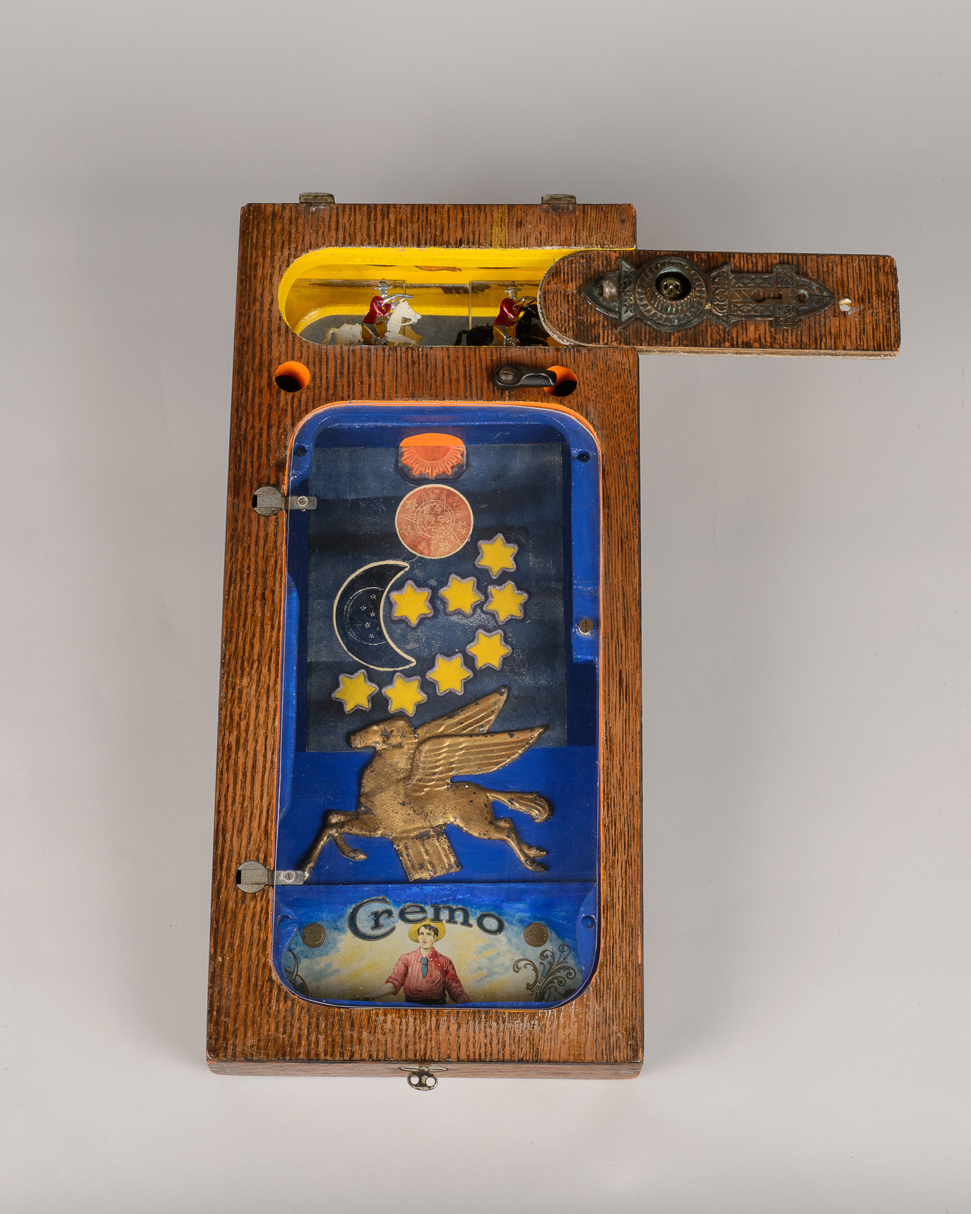 Assemblage on wood with a blue inlay design with a flying horse in gold, crescent moon and eight yellow tars, metal knobs and holes, and a vintage metal piece that says "Cremo" on it.