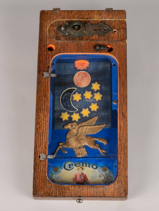 Assemblage on wood with a blue inlay design with a flying horse in gold, crescent moon and eight yellow tars, metal knobs and holes, and a vintage metal piece that says "Cremo" on it