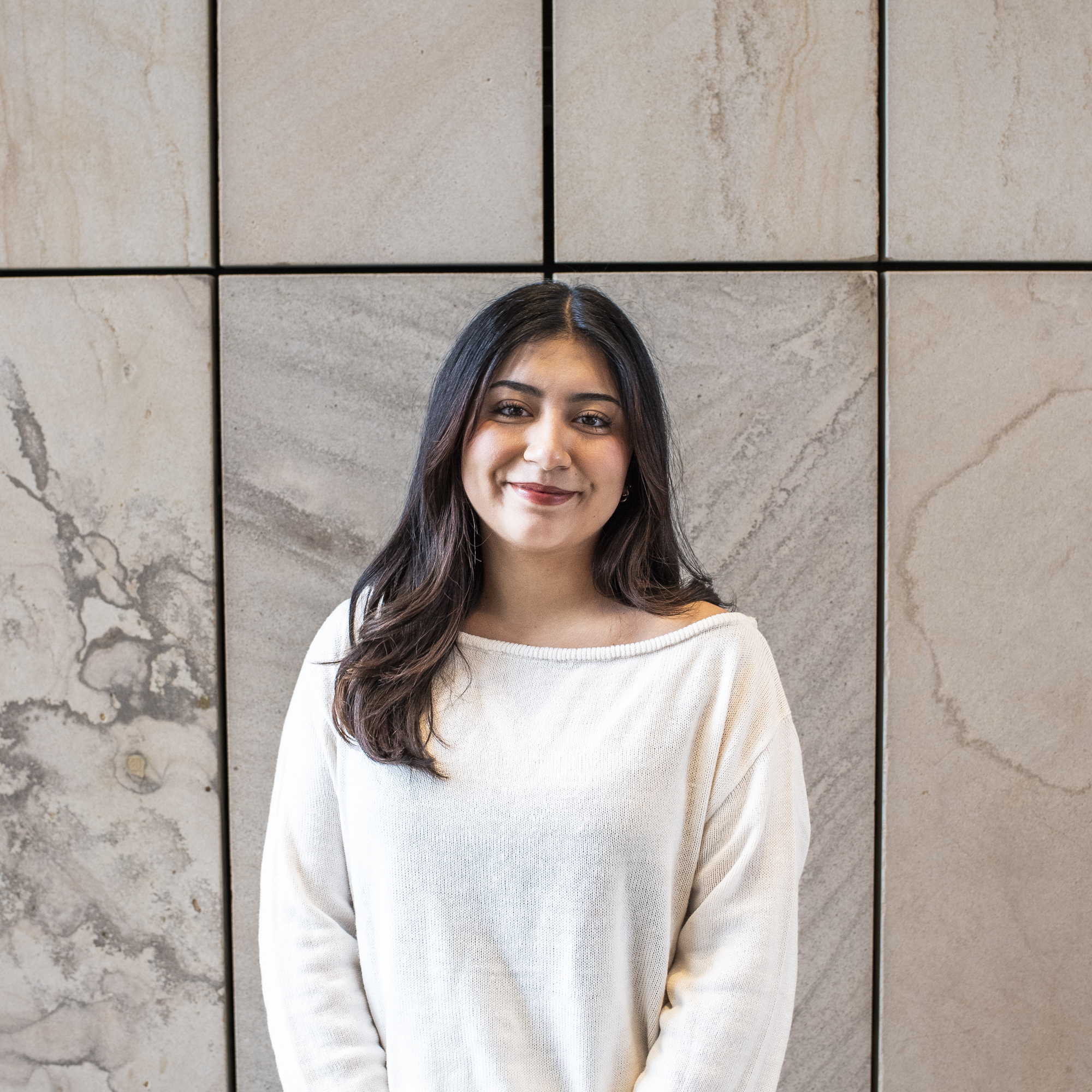 Woman with long brown hair in a white sweater standing in front of a wall made of sandstone slabs
