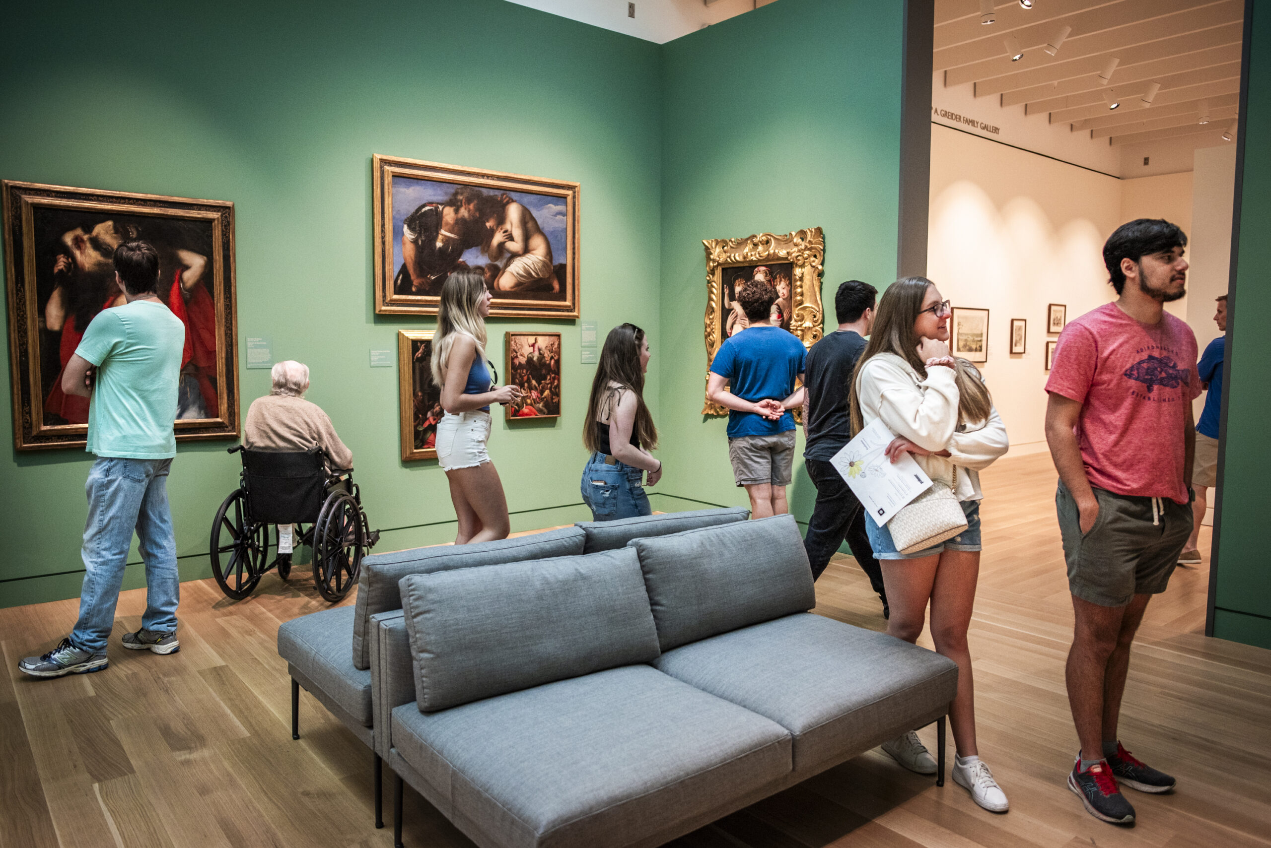 Varied group of visitors to an art museum with green walls and Baroque paintings around a grey couch in the center of the room