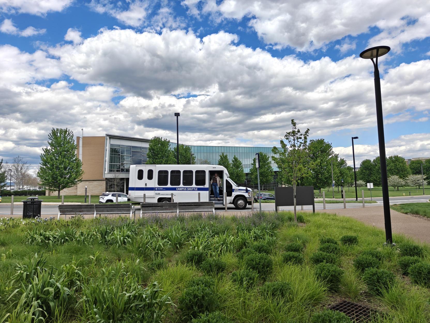 White shuttle bus stopping on a road with grass in the foreground, a building behind it, and a blue sky with white clouds above