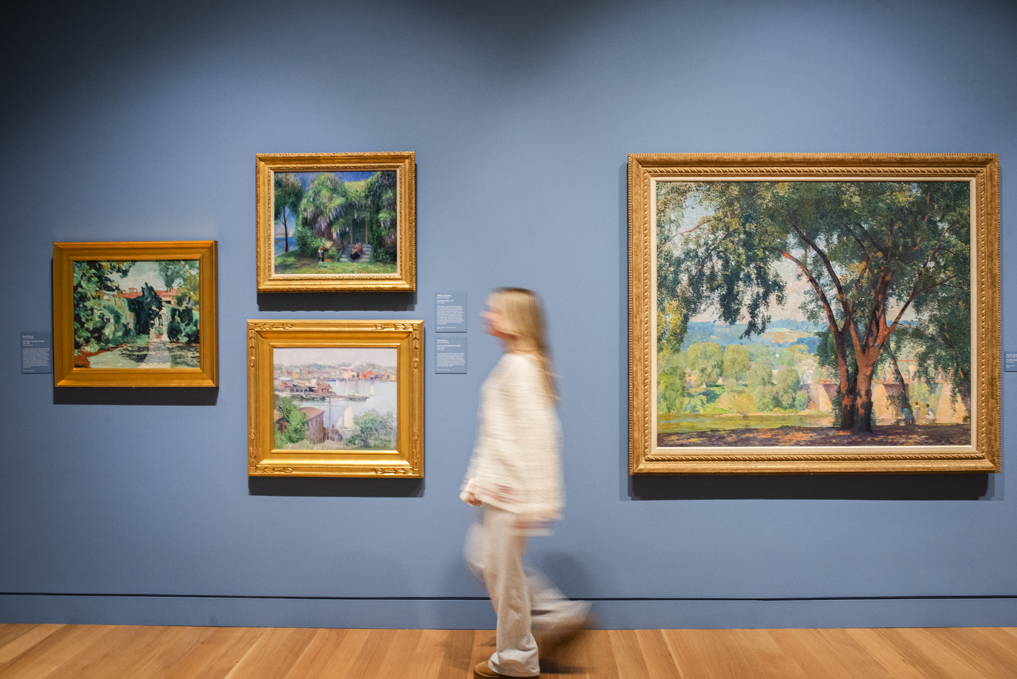 A blurred figure in white is in motion as they walk along a blue wall with Impressionist paintings of leaves and landscapes in an art gallery.