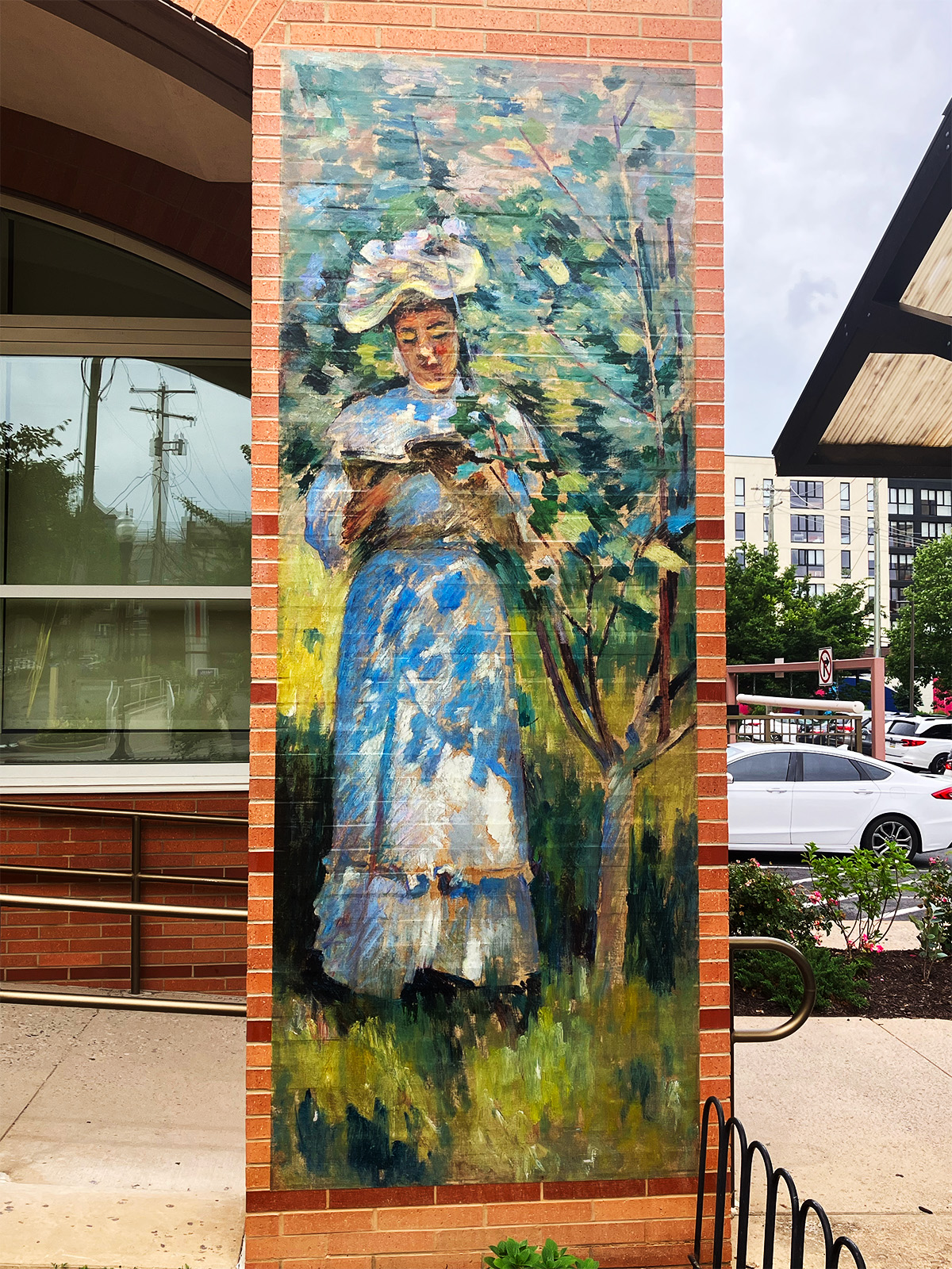 Mural of an artwork depicting a standing woman in frilly Victorian clothes and sunhat reading beneath the shade of a dappled tree. The mural is on the brick column of a building and has a parking lot at right.
