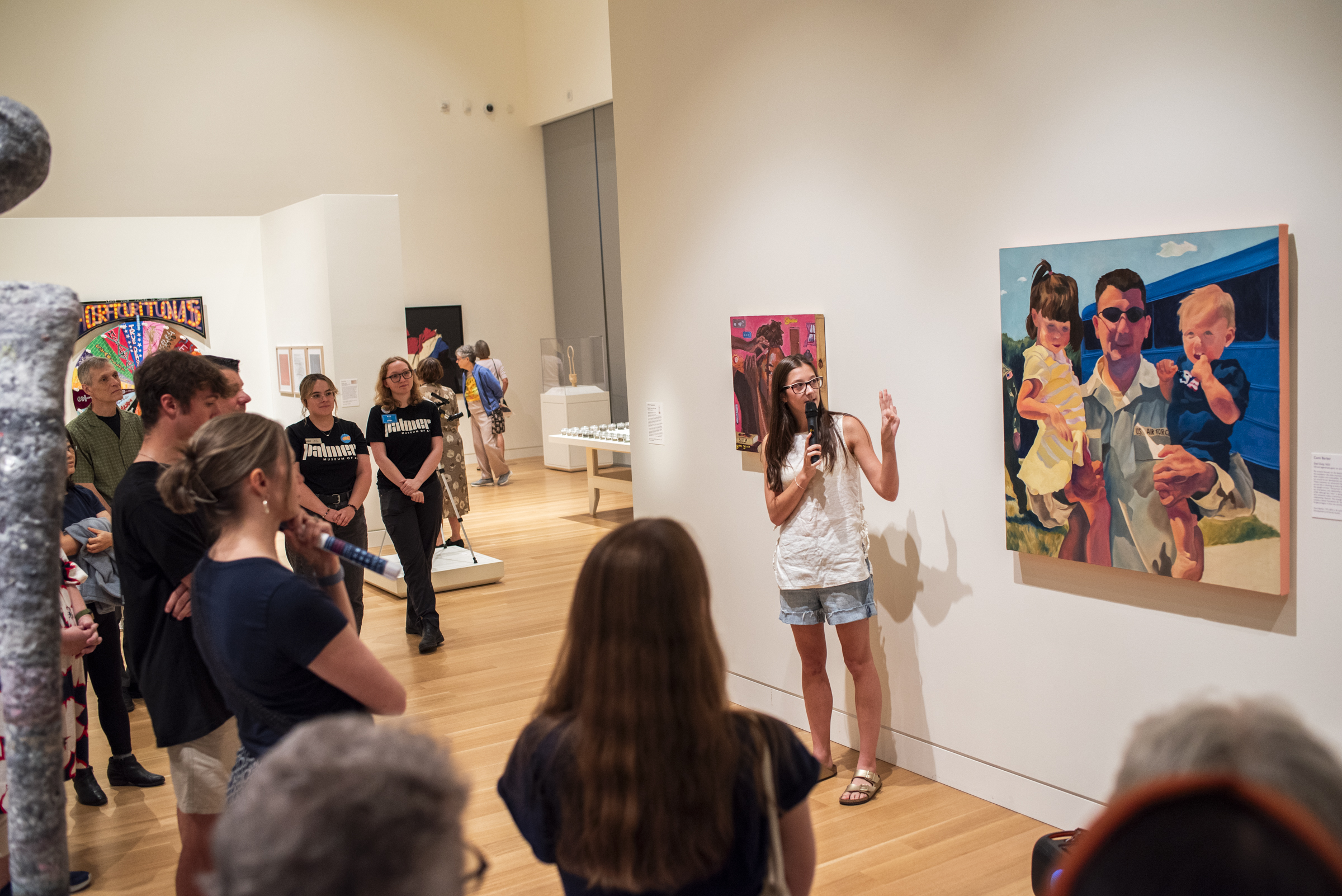 A group in a gallery stands around a person talking about their artwork as it hangs on the wall.