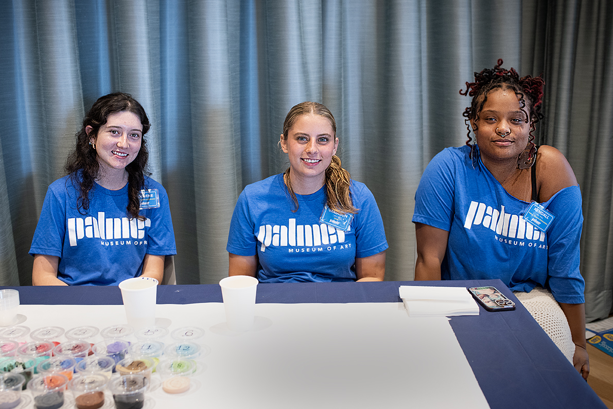 Three students in matching blue Palmer Museum t-shirts sit at a table with art supplies in front of a curtain.