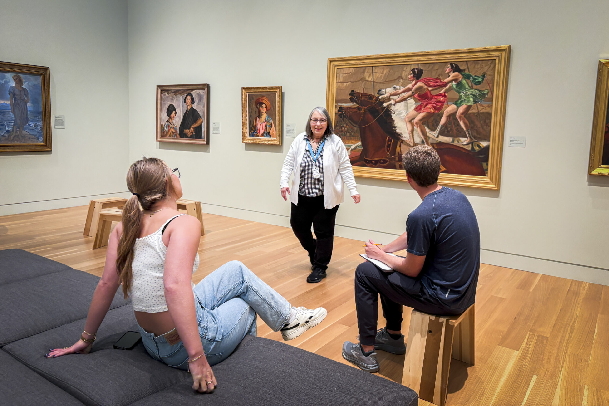 Two young people sit in an art gallery on a couch while listening to another person who is standing and talking to them.