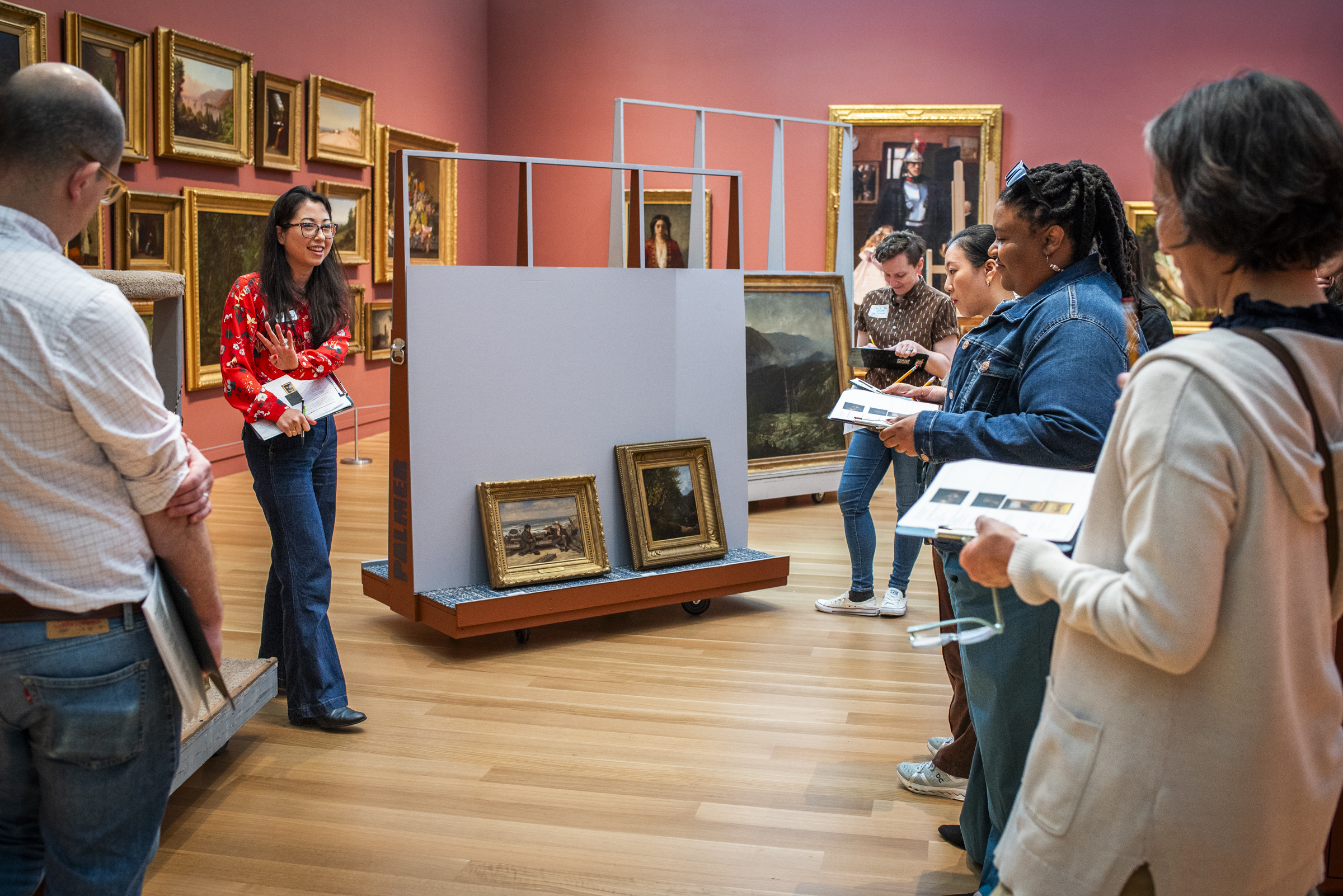 A group of faculty surround a curator in a red-walled gallery with artworks on carts.