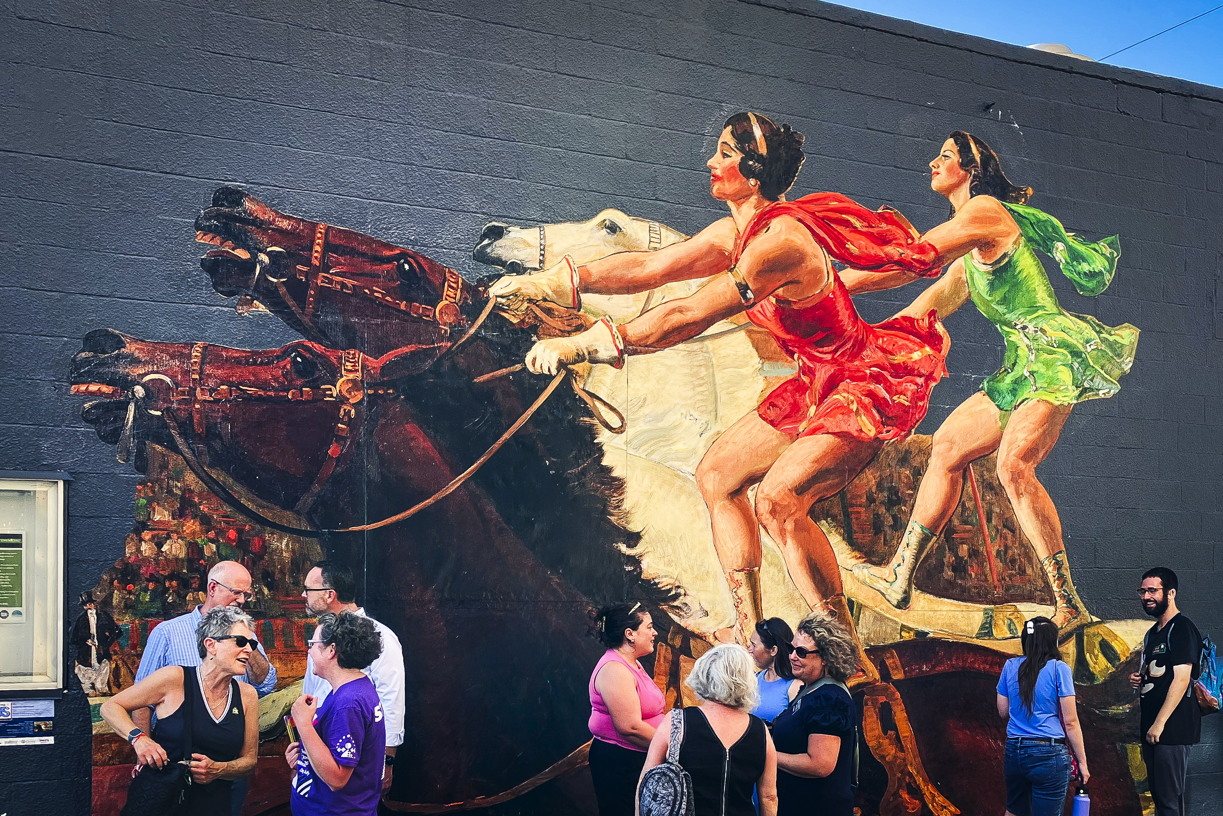 A group of people stand in front of a mural depicting two women in green and red standing and riding on brown and white horses.