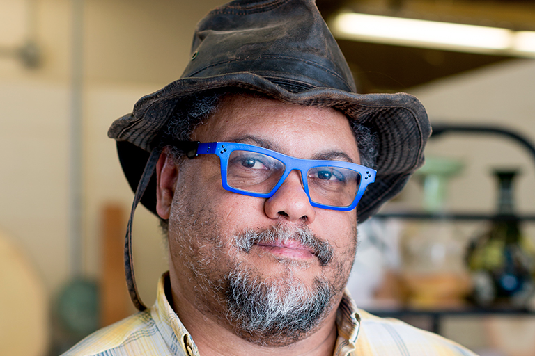 Artist Malcolm Mobutu Smith, a Black man with a salt and pepper goatee wearing bright blue glasses, a worn brown brimmed hat.