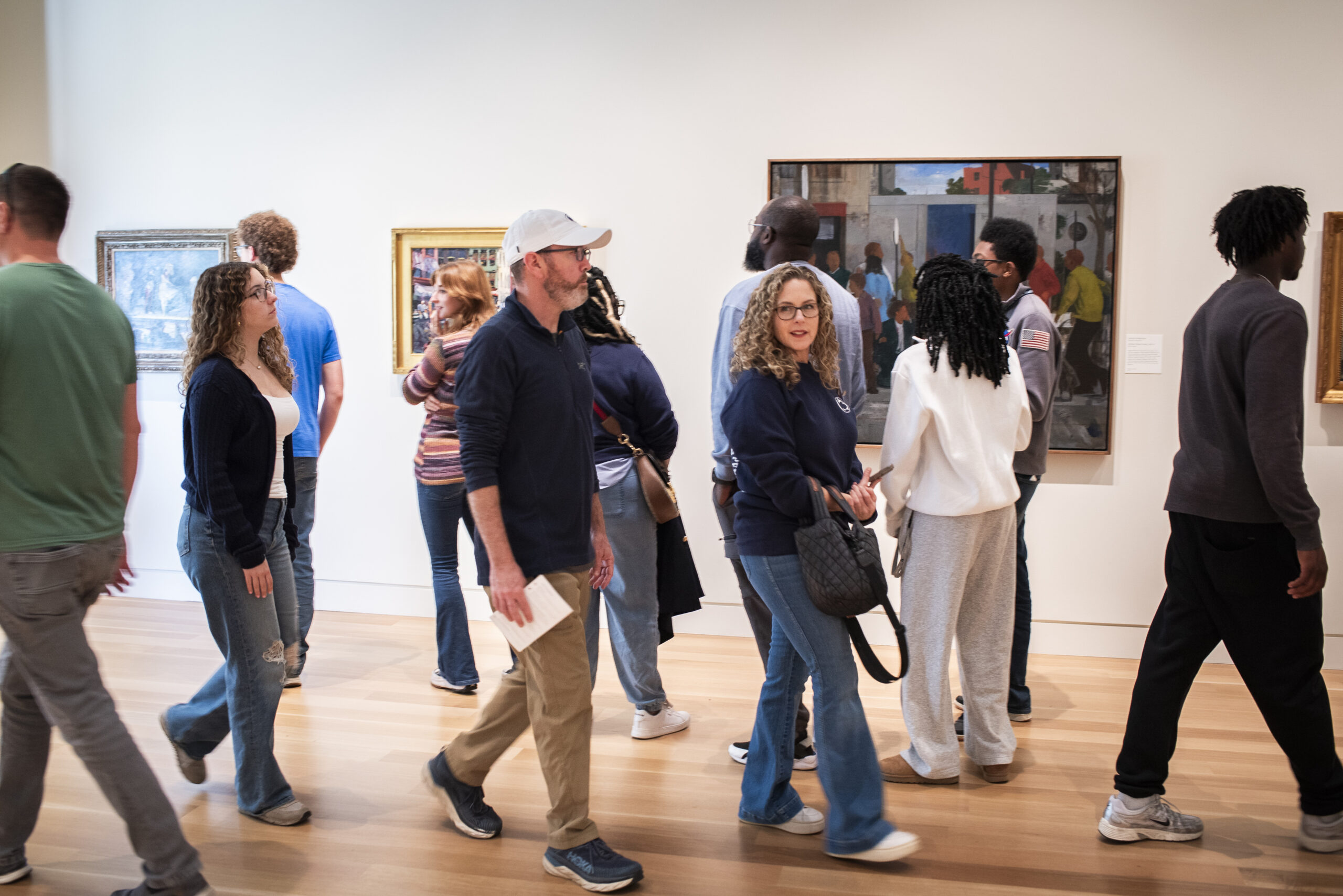 A large group of people of varied ages, backgrounds, ethnicities, and genders, walk through an art gallery with art on the walls.