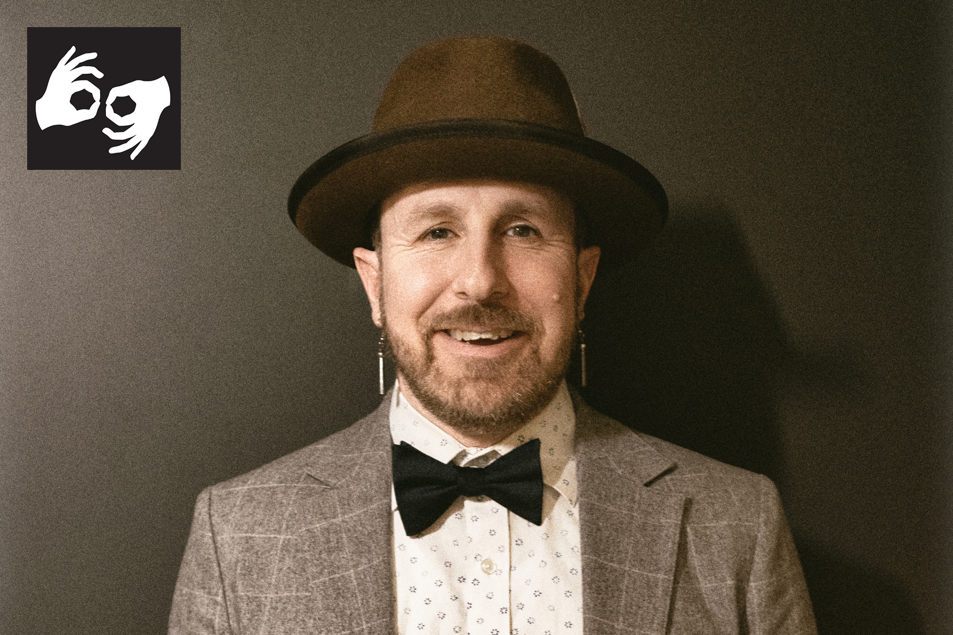 Artist Ryan RedCorn in a brimmed hat, dark bowtie, and plaid jacket, smiling at the camera. He wears long earrings. A black and white ASL interpretation icon with two hands signing sits at top left corner.