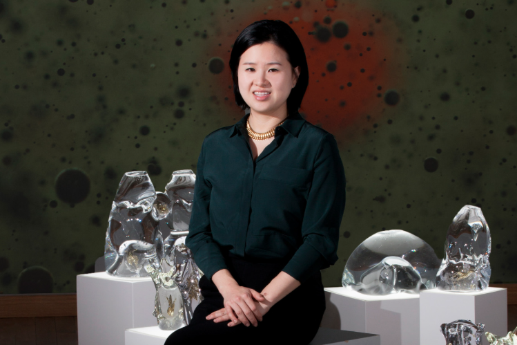 Artist Wonjung Choi sits amid clear class sculptures on pedestals while she smiles at the camera in a dark button down shirt.